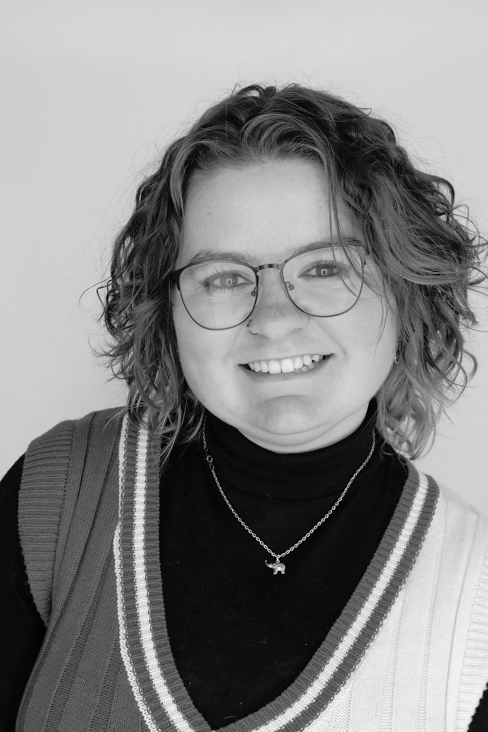 Black-and-white portrait of a smiling person with curly hair, glasses, and a necklace, against a plain background.