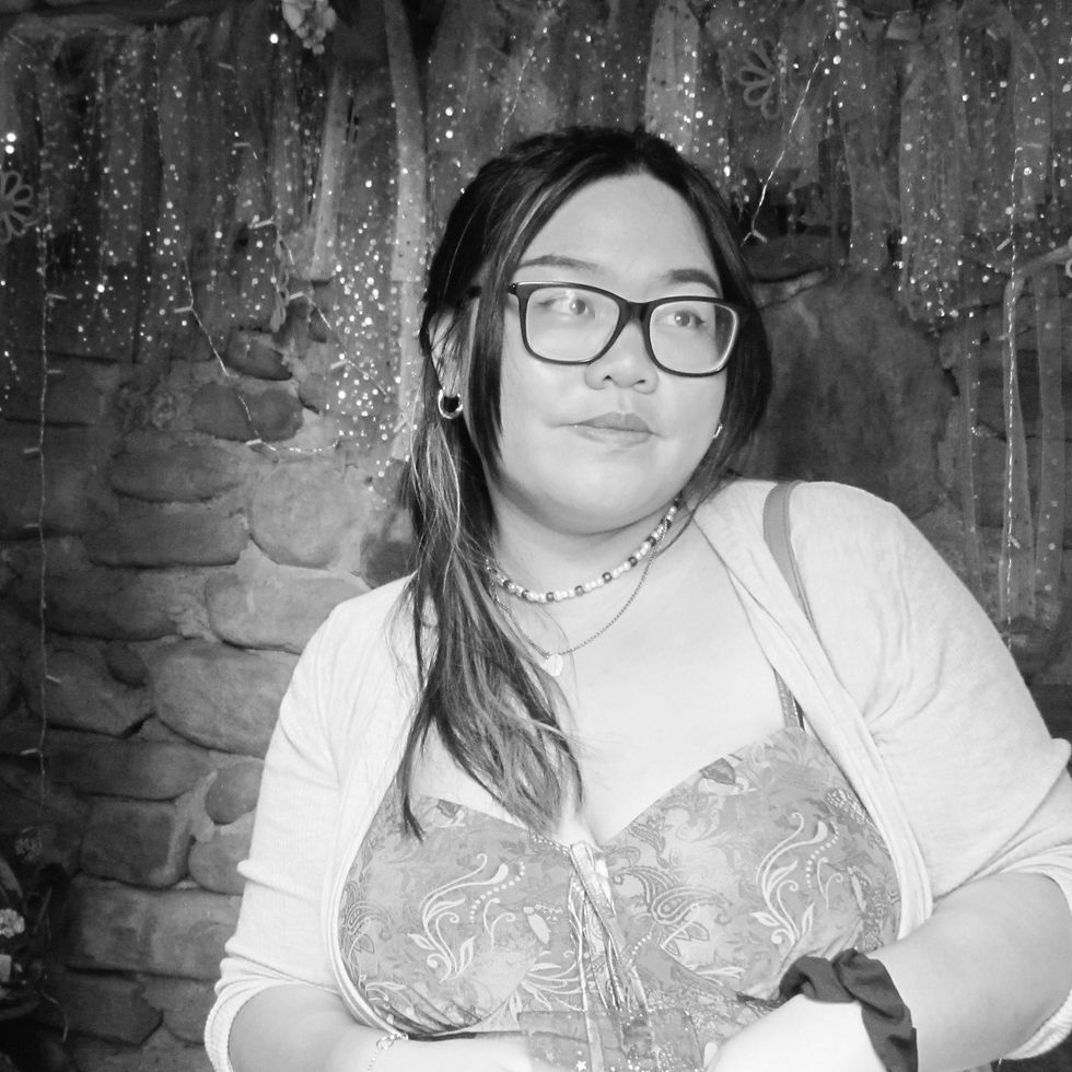 Woman with glasses stands in front of a stone wall adorned with glittery fabric. She's wearing a patterned dress, looking thoughtful.
