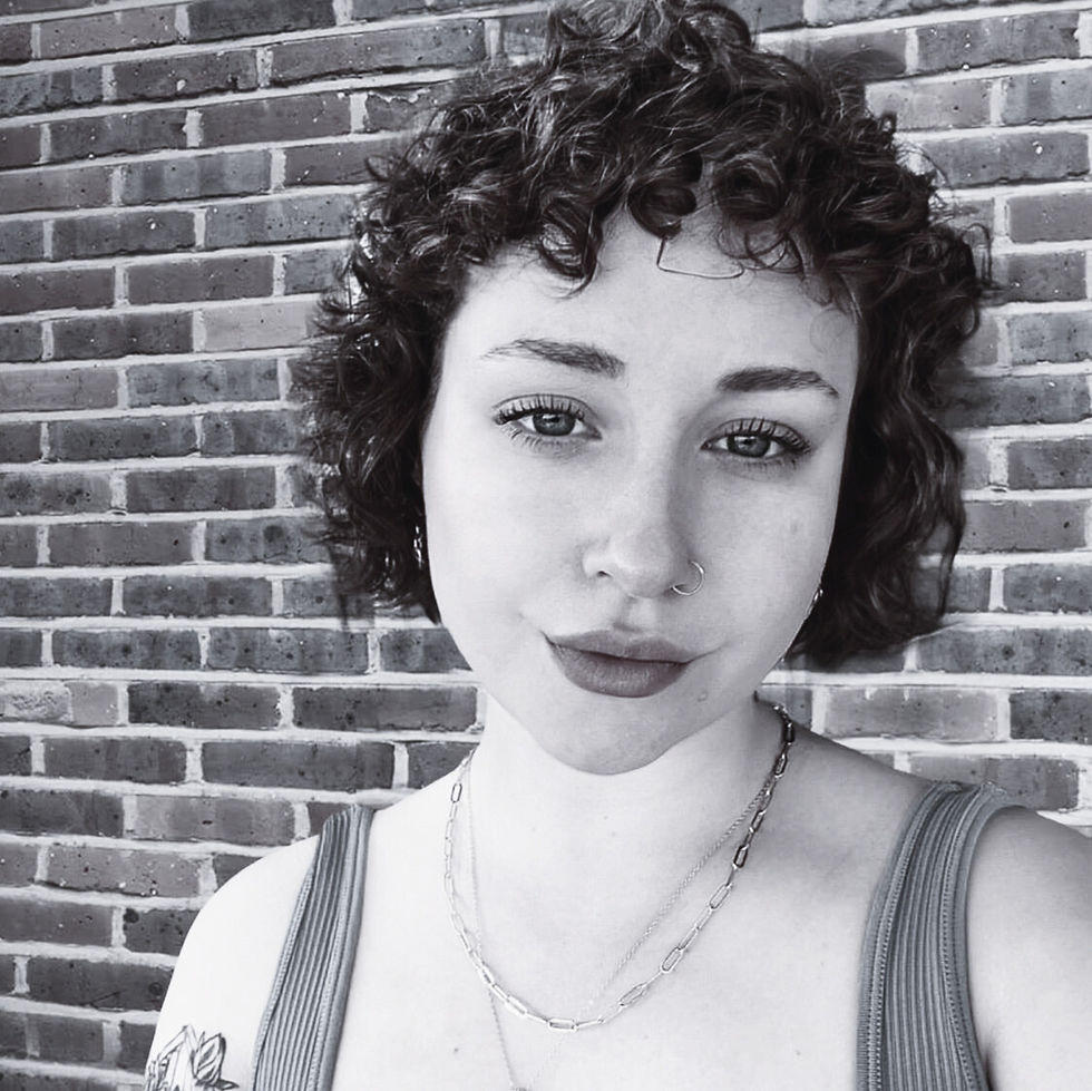 Woman with curly hair and a nose ring poses against a brick wall, wearing a chain necklace and tank top. Black and white photo.