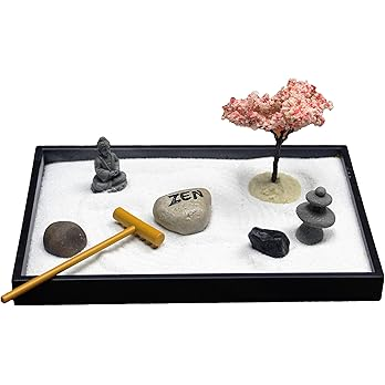 Zen garden tray with white sand, a small statue of Buddha, a miniature cherry blossom tree, a wooden rake, three stacked balancing stones, and three different sized decorative rocks, including one engraved with the word “ZEN.” The arrangement promotes relaxation and mindfulness.