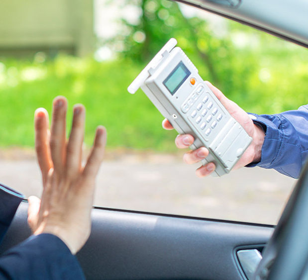A imagem mostra o volante de um carro e a mão de um homem, negando o aparelho de bafômetro apresentado pela mão de outro homem,