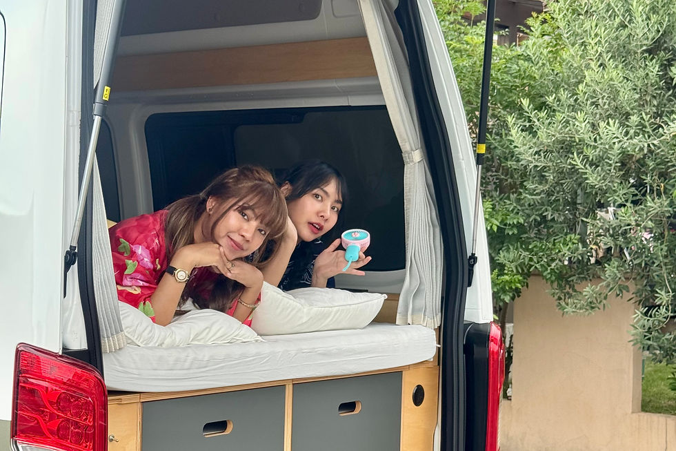 Two women relax inside a campervan's open trunk, one holding a small fan. The setting is outdoors with green foliage visible. They appear content. Japan campervan, Tokyo campervan, Tokyo, vanlife, Japan road trip, camper van, summer tokyo, summer japan