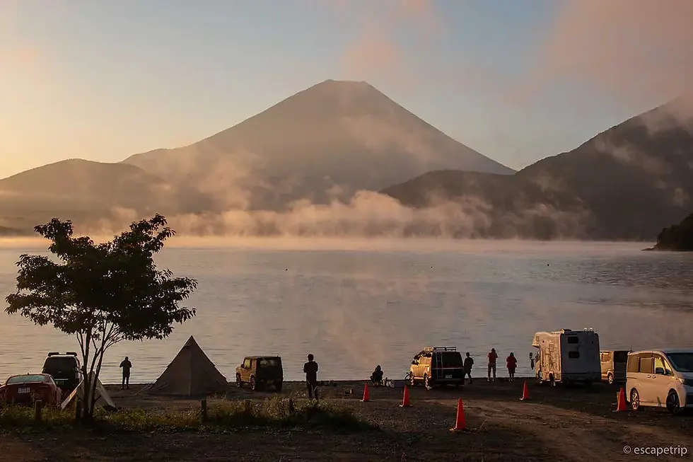 Camping scene by a misty lake at sunrise, with Mount Fuji in the background. People stand near vehicles and tents, creating a serene mood.