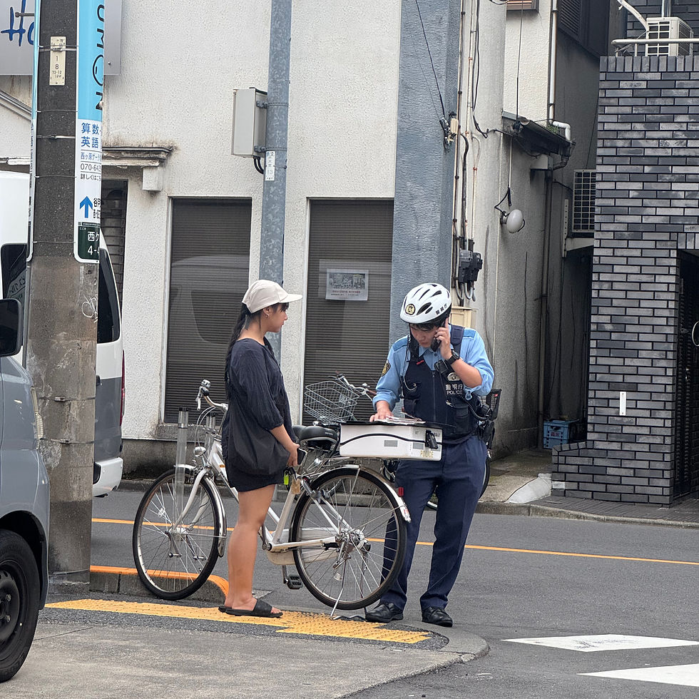 A police officer talks on the phone, writing on a clipboard by bicycles. A person in a hat stands beside them on a city street corner.