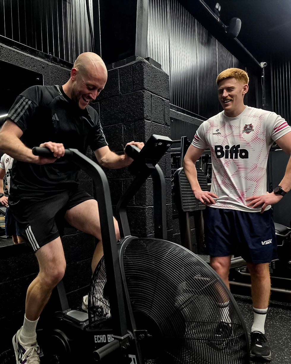 Two men in a gym; one exercising on a bike, smiling, wearing black. The other watches, smiling, in a white Biffa jersey. Dark interior.