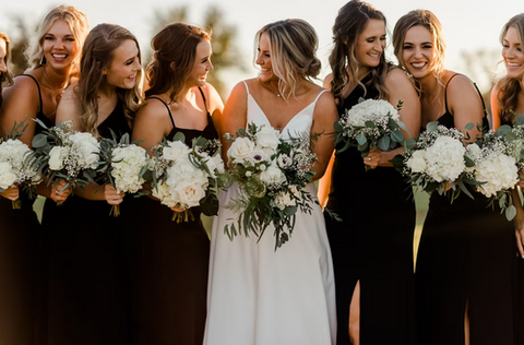 Bridesmaids holding white florals in black dresses with bride and custom florals designed and executed by all-inclusive wedding planners.