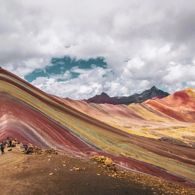 Strati ondulati di terra in tonalità arancio, porpora e giallo si estendono sotto un cielo nuvoloso a Vinicunca, una meta da non perdere per gli amanti del trekking in un tour in Perù