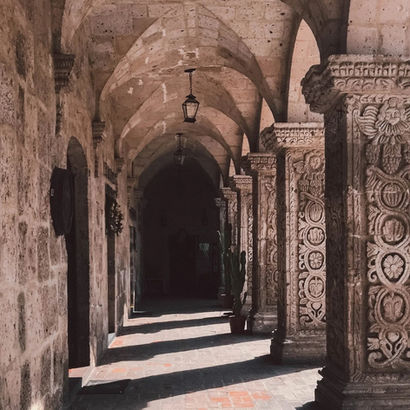 Un corridoio ad arco con soffitti a volta, sorretto da colonne ornate con intagli complessi. La luce solare calda filtra attraverso, proiettando ombre sul pavimento in pietra, creando un'atmosfera di eleganza storica