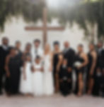 Bride and groom standing at the center with their wedding party, dressed in black and white attire, posed in front of a cross