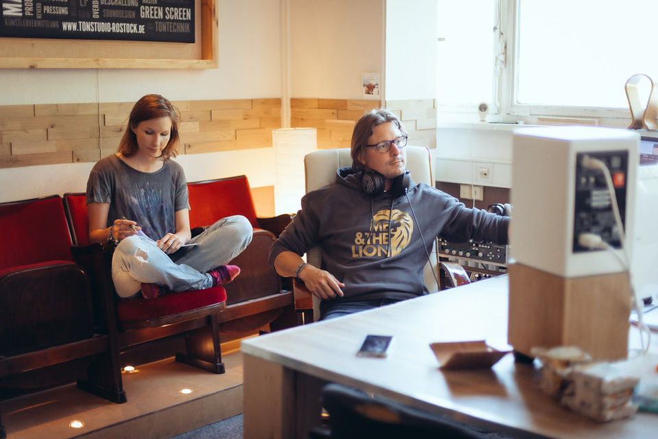Foto einer Songwriterin und eines Produzenten im Tonstudio. Er sitzt entspannt am Studio-Computer, während sie etwas dahinter im Schneidersitz auf gemütlichen Theatersesseln sitzt und konzentriert ihre Sheets liest.