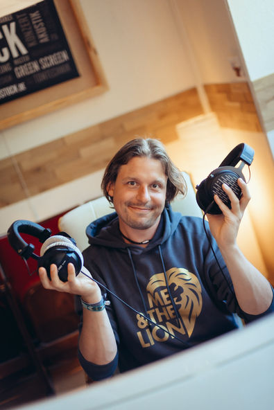 Foto des Musikproduzenten Leo Sieg in der Studio-Regie. Er sitzt vor dem Mischpult, hält zwei unterschiedliche Studiokopfhörer in den Händen und blickt lächelnd in die Kamera.