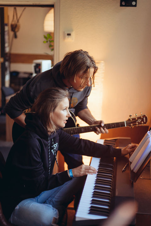 Foto von zwei jungen Musikern beim Songwriting im Tonstudio. Er hält eine Akustikgitarre und beugt sich über das Klavier, während sie sitzend Klavier spielt. Beide schauen auf ein Notizbuch. Gedämmtes Licht sorgt für eine gemütliche Studioatmosphäre.