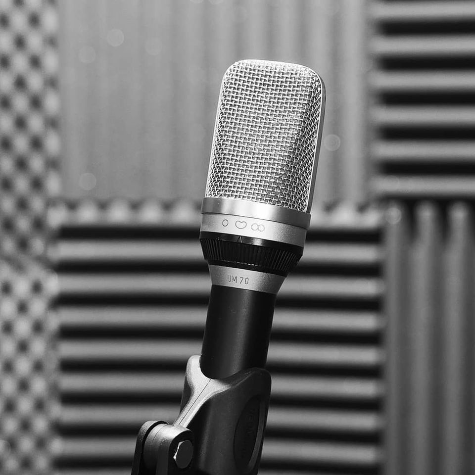 Schwarzweißfoto des Großmembran-Mikrofons UM70 von Neumann/Gefell im Vordergrund. Im unscharfen Hintergrund ist eine Vocal Booth eines Tonstudios zu sehen.