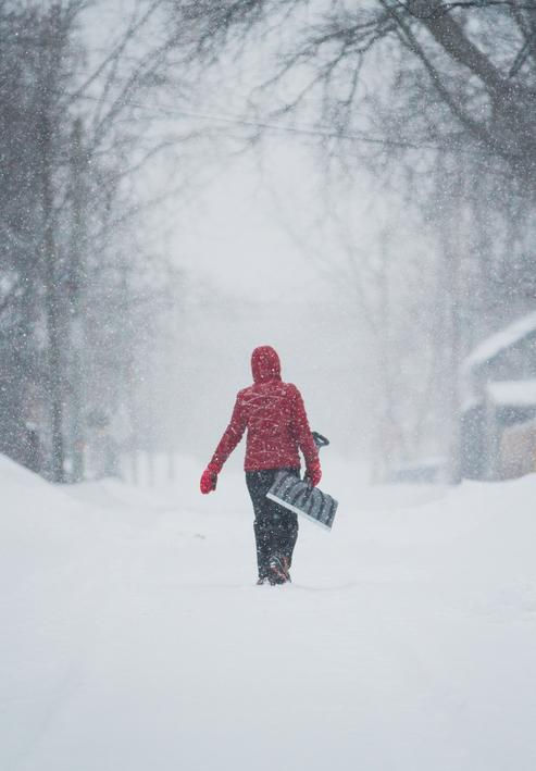Person walking down a snowy street carrying a shovel during winter weather.