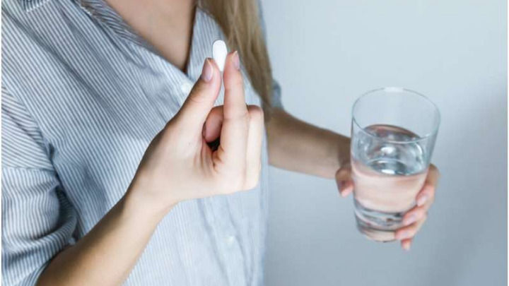 Una mujer con camisa a rayas sostiene una pequeña pastilla blanca entre sus dedos mientras tiene un vaso de agua en la otra mano, preparándose para tomar su medicación.