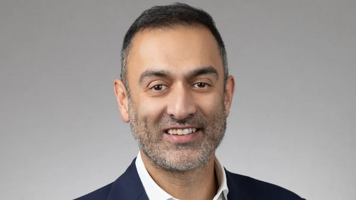 Retrato de un hombre de mediana edad con barba corta, sonriendo con un traje azul y camisa blanca sobre un fondo gris neutro.