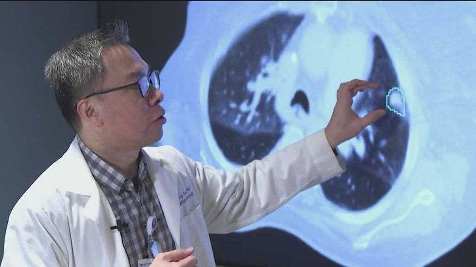 An Asian doctor with glasses points at a large screen displaying a lung CT scan with a highlighted area. He appears to be explaining medical findings or a condition.