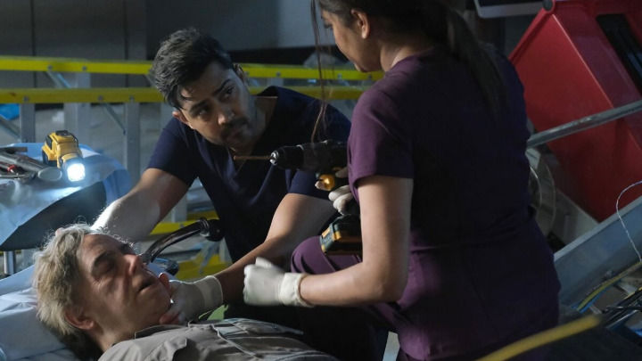 In a dimly lit emergency setting, two medical professionals in dark scrubs urgently treat an unconscious patient. They use a power drill and other tools, maintaining intense and focused expressions during the procedure.