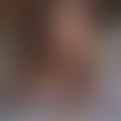 This image shows a front-facing close-up of a young woman with long, wavy blonde hair. She is sitting on a tufted sofa with vertical stripes, wearing a light blue shirt and a thin chain around her neck. Her expression is serious and directed toward the camera, with soft lighting highlighting her features. To her left, a blue-gray blanket is visible, creating a calm and somewhat melancholic atmosphere.