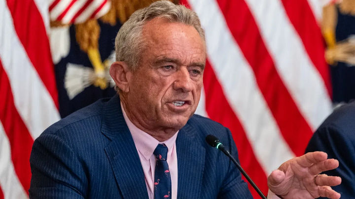 Robert F. Kennedy Jr. is speaking at a table, gesturing with his hand. He is wearing a pinstripe suit and a pink tie with flamingos. American flags are in the background.