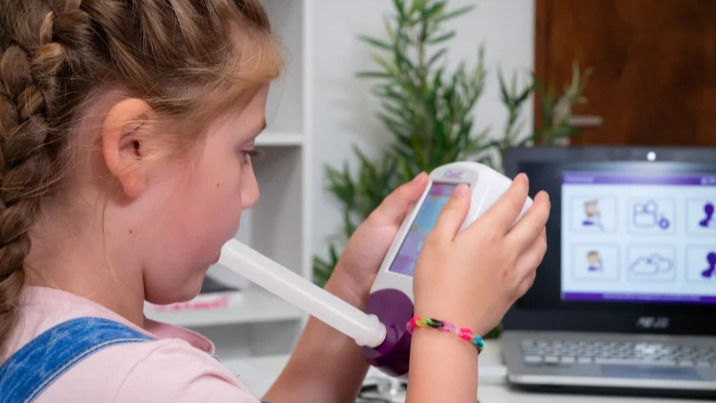 A young girl with braided hair uses a handheld spirometer device for a lung function test, while a laptop in the background displays medical icons and data in a bright clinical room.