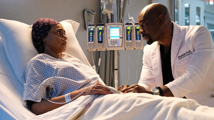 In a brightly lit clinical room, a male doctor in a white coat sits by a patient's bed, holding her hand. They share an emotional and compassionate moment while maintaining serious, focused, and concerned expressions.
