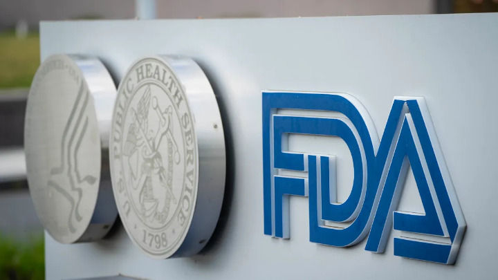 Close-up of the FDA logo in 3D blue letters on a white outdoor sign. Metallic circular seals of the U.S. Department of Health and Human Services are visible to the left.