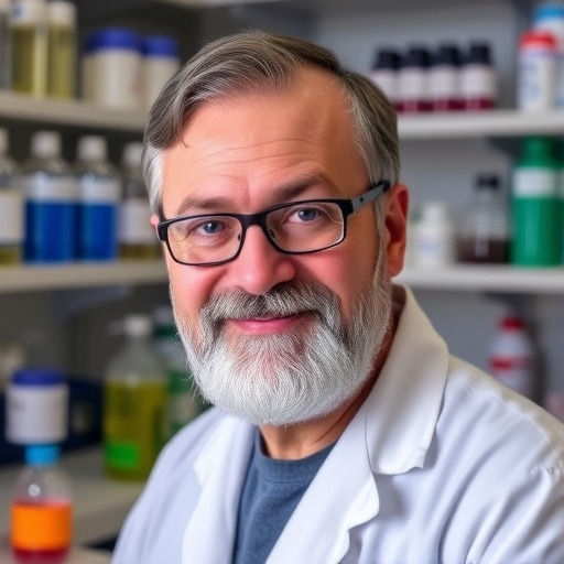 Un científico de barba blanca y gafas sonríe a la cámara en un laboratorio, vestido con una bata blanca, con estantes llenos de frascos químicos desenfocados detrás de él.