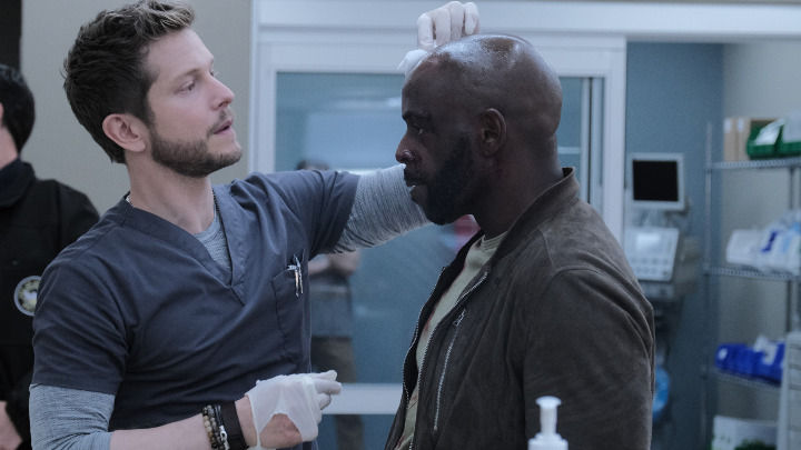 Un médico con uniforme gris y guantes trata cuidadosamente una herida en la cabeza de un paciente con chaqueta marrón. Están en una sala clínica con suministros médicos visibles sobre un mostrador cercano.