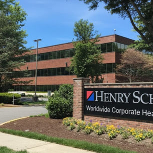 The Henry Schein Worldwide Corporate Headquarters sign sits next to a curving road. The brick office building is surrounded by lush green trees under a blue sky.