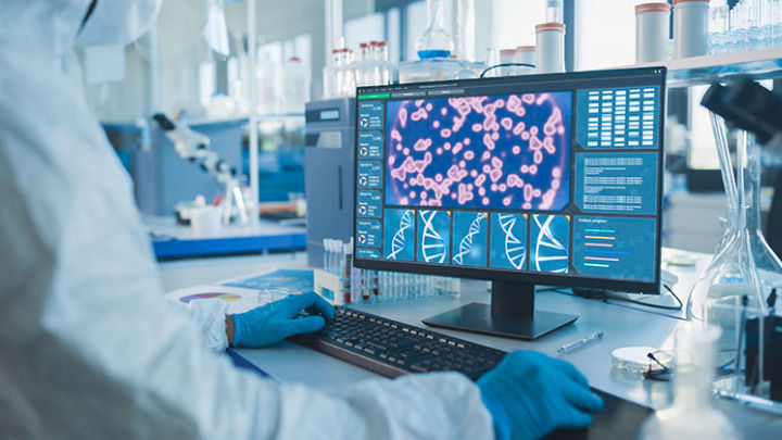 A researcher in protective gear and blue gloves works on a computer displaying cell structures and DNA models in a brightly lit, modern medical laboratory setting.