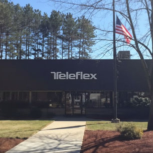 The Teleflex corporate building is a dark, rectangular structure with the white logo, set in a wooded area with a US flag flying on a pole in the front.