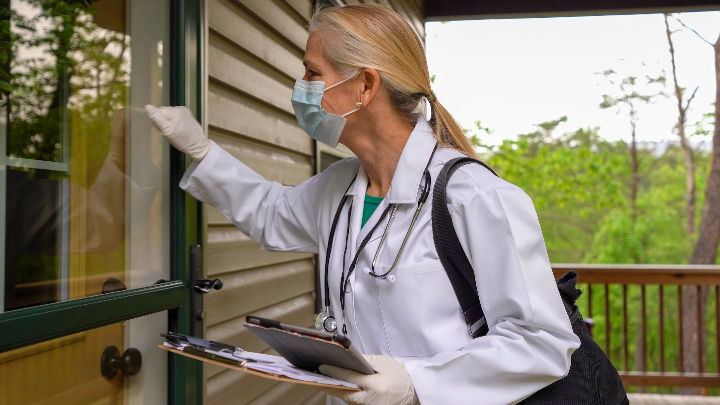 Masked doctor in white coat knocks on a door while standing on a porch, holding a clipboard. Background shows green trees and nature.