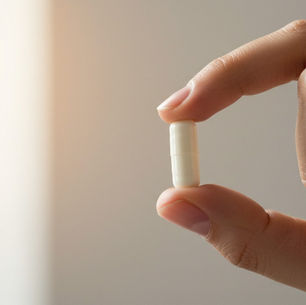 La imagen muestra una mano sosteniendo una pastilla blanca y alargada con el pulgar y el índice. La toma es de cerca, con un fondo borroso y una luz cálida que entra por la izquierda.