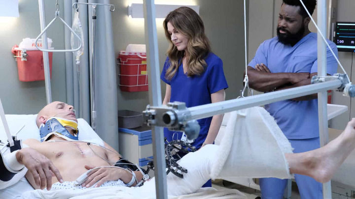 In a hospital room, a female and a male doctor from The Resident observe a patient lying in bed with a neck brace and an elevated leg. The scene conveys a serious medical atmosphere.