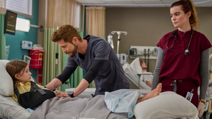 A male doctor in blue scrubs comforts a young girl with an arm sling in a hospital room, while a female nurse in burgundy scrubs watches them from the bedside.