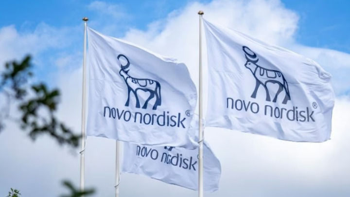 Tres banderas blancas con el logotipo azul de Novo Nordisk y el icónico símbolo del buey Apis ondean bajo un cielo brillante y nublado, representando la identidad corporativa de la farmacéutica.