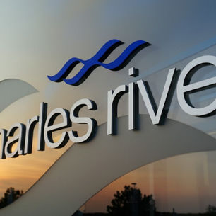 A close-up of the Charles River logo and wordmark, adhered to a reflective glass building. The glass reflects an orange sunset and surrounding trees.