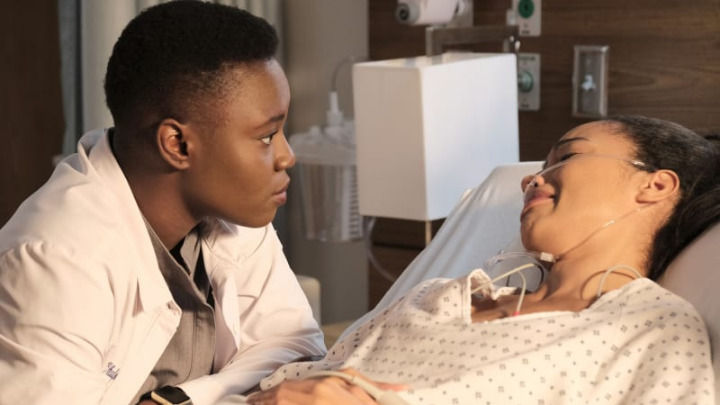In this scene from The Resident, Mina Okafor leans in closely to comfort a patient lying in a hospital bed. The intimate lighting emphasizes the emotional connection and care provided during this quiet medical moment.
