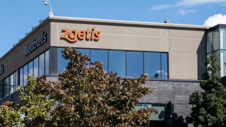 La imagen muestra un edificio de oficinas de Zoetis. El logo naranja de la empresa está en la fachada, con grandes ventanas oscuras y árboles en primer plano bajo un cielo azul.
