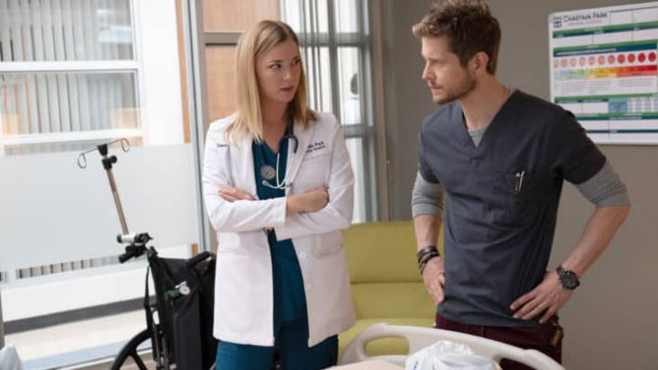 Dos profesionales médicos concentrados, una mujer con bata blanca y un hombre con uniforme oscuro, están junto a la cama de un paciente y mantienen una seria discusión clínica mientras se miran el uno al otro.