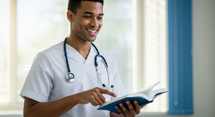 Smiling person in a white coat with a stethoscope reads a book, pointing at a page. Bright room with blue blinds in the background.