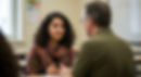 A young woman with dark curly hair and a colorful scarf gestures while speaking to a man in glasses and an olive green shirt, seated in a classroom, possibly during a tutoring session or conversation.
