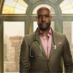 This image shows an African American man with a beard, wearing a sharp brown suit, pink shirt, and a stethoscope around his neck. He stands before a large window overlooking a city skyline.