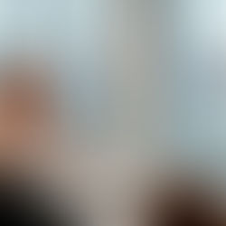 A young redhead woman in a white shirt and dark pants gestures while presenting to a group of students in a modern classroom with large windows overlooking urban buildings.