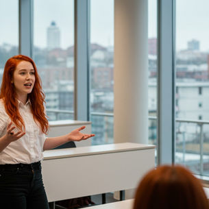 A young redhead woman in a white shirt and dark pants gestures while presenting to a group of students in a modern classroom with large windows overlooking urban buildings.