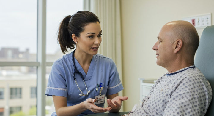 A nurse practitioner in blue scrubs talks to a patient in a hospital gown. They appear engaged in conversation in a bright hospital room.
