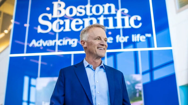 A smiling, middle-aged man in a blue suit and light-colored shirt stands confidently. Behind him is a large blue Boston Scientific backdrop featuring the company name and the slogan "Advancing science for life."