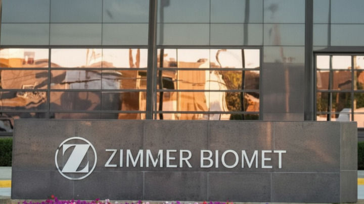 A dark grey stone sign displaying the white Zimmer Biomet logo and text sits in front of a building with reflective windows, over a bed of purple flowers.