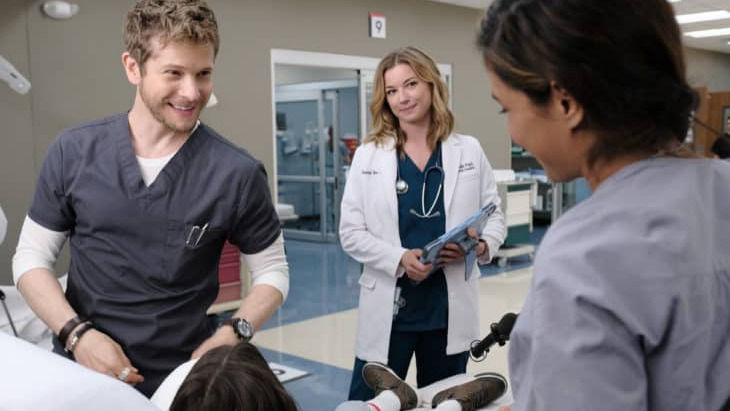 In a bright hospital room, three medical professionals from The Resident smile warmly at a young patient lying in bed. The scene captures a moment of compassionate care and teamwork within the clinical setting.